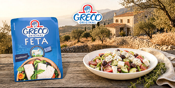 Verpackung von Greco Original Feta neben einer Schüssel mit griechischem Bauernsalat auf einem Holztisch vor mediterraner Landschaft.